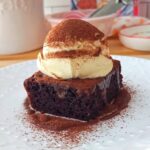 Slice of chocolate cake topped with ganache and mascarpone cream on a white plated. Dusted with cocoa powder